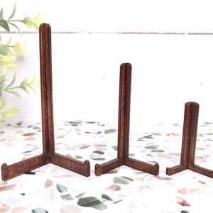 May include: Three dark brown wooden display stands of varying heights, each featuring a vertical support post with a central slot and two horizontal feet extending forward to hold items securely in an upright position.