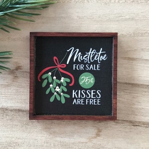 May include: A black framed sign with a white background and red and green illustrations of mistletoe. The sign reads "Mistletoe for Sale 25¢ Kisses are Free".