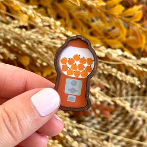 Peut inclure: Épinglette en bois en forme de distributeur de boules de gomme, remplie de boules de gomme orange en forme de citrouille. L'épinglette a une bordure marron et un corps orange clair. Le fond est un mélange de plantes séchées jaunes et marron.