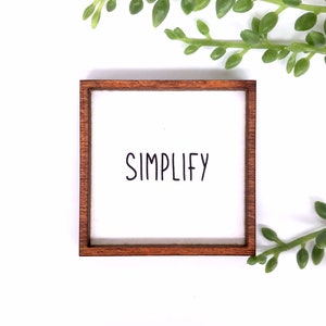 May include: A wooden framed sign with the word "SIMPLIFY" written in black script on a white background.