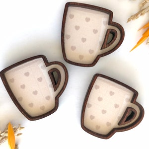 May include: Three wooden coffee mugs with a heart pattern. The mugs are brown with a light brown interior and a white heart pattern.
