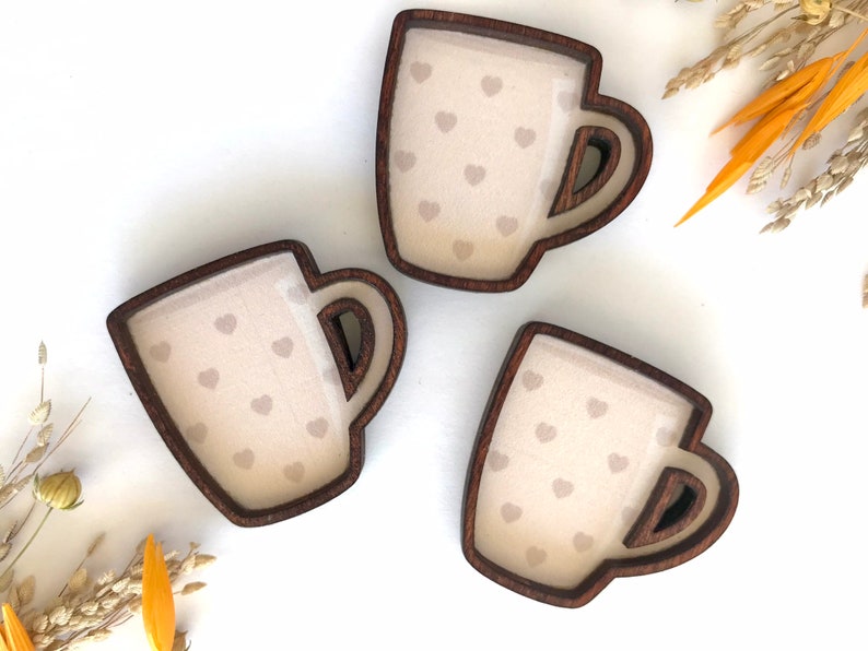 May include: Three wooden coffee mugs with a heart pattern. The mugs are brown with a light brown interior and a white heart pattern.