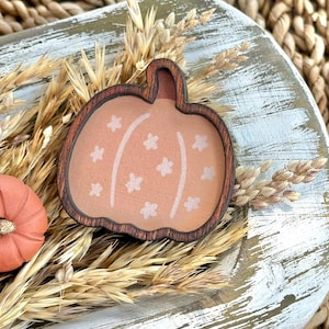 Peut inclure: Décoration en forme de citrouille en bois, avec un centre orange clair et des accents d'étoiles blanches. La citrouille est entourée d'épis de blé et repose sur un plateau en bois vieilli, évoquant le thème de l'automne.