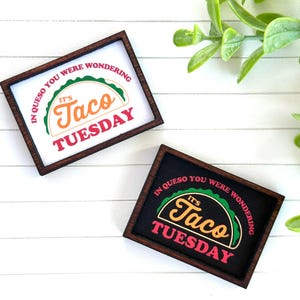 May include: Two small, rectangular wooden signs with the text "IN QUESO YOU WERE WONDERING, IT'S TACO TUESDAY." One sign has a white background, the other black. The text is in green, orange, and red.