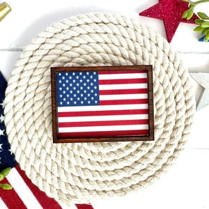 May include: A small wooden frame with an American flag design. The frame is sitting on a white rope mat. There are red, white, and blue stars and other decorations around the frame.