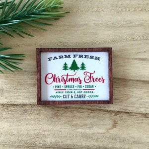 May include: A rectangular wooden sign with the text "FARM FRESH Christmas Trees" in red and white. The sign also includes the text "PINE &bull; SPRUCE &bull; FIR &bull; CEDAR" and "APPLE CIDER & HOT COCOA". A sprig of evergreen is in the background.