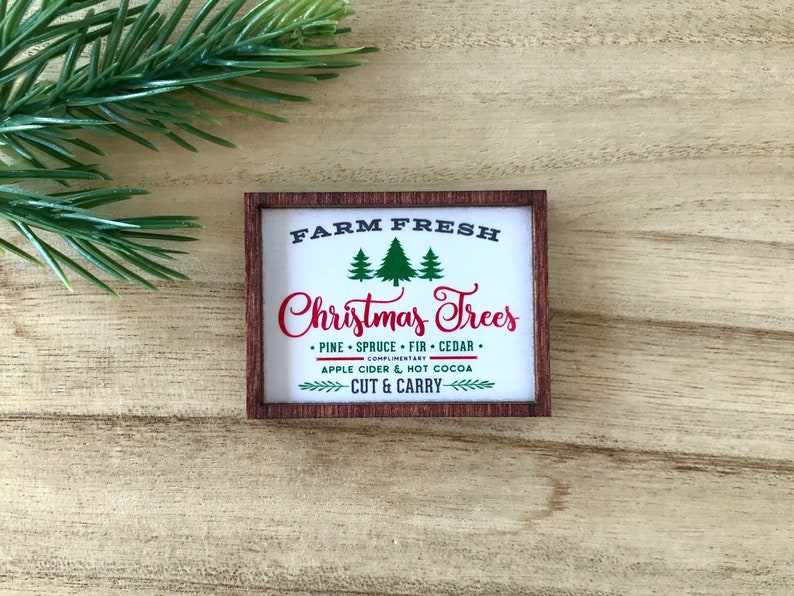 May include: A rectangular wooden sign with the text "FARM FRESH Christmas Trees" in red and white. The sign also includes the text "PINE &bull; SPRUCE &bull; FIR &bull; CEDAR" and "APPLE CIDER & HOT COCOA". A sprig of evergreen is in the background.