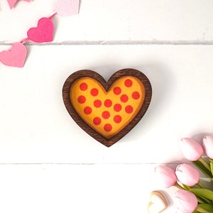 May include: A wooden heart-shaped dish with a pizza design. The dish is painted yellow with red pepperoni circles. The dish is on a white wooden background.