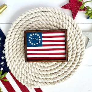 May include: A framed print of the American flag with the date 1776 in the upper left corner. The frame is dark brown wood and the print is on a white background. The print is sitting on a white rope mat.