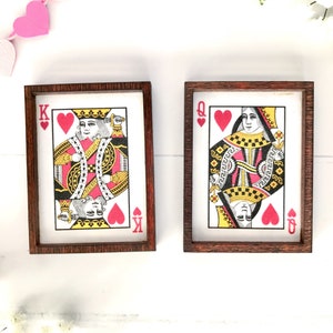 May include: Two framed playing card prints featuring the King and Queen of Hearts. The cards have a classic design with red, black, and gold details, set in dark brown wooden frames. A decorative item for a game room or home decor.
