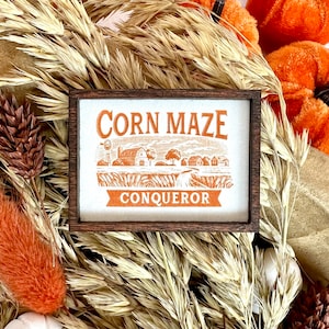 May include: A wooden sign with the text "CORN MAZE CONQUEROR" in orange lettering. The sign features a small illustration of a farmhouse and cornfield.
