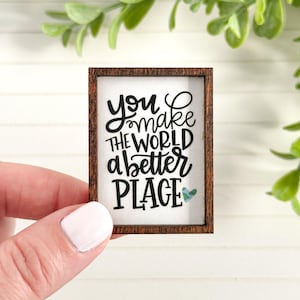 May include: Small rectangular wooden sign with the phrase "you make the world a better PLACE" in black script. The sign has a dark brown frame and a small teal heart. The sign is held by a person.