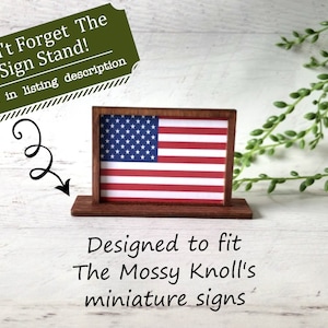 May include: A miniature wooden sign stand displaying an American flag. The sign is red, white, and blue. The text on the image says, "Don't Forget The Sign Stand!" and "Designed to fit The Mossy Knoll's miniature signs."