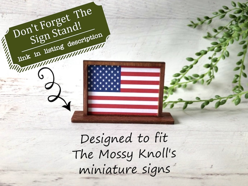 May include: A miniature wooden sign stand displaying an American flag. The sign is red, white, and blue. The text on the image says, "Don't Forget The Sign Stand!" and "Designed to fit The Mossy Knoll's miniature signs."