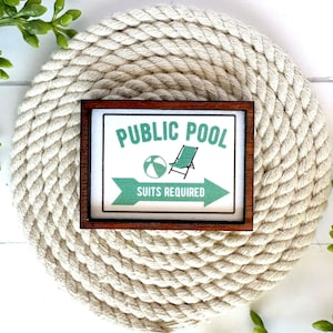 May include: A wooden sign with the text "PUBLIC POOL SUITS REQUIRED" in white lettering. The sign has a green beach ball and a green beach chair graphic. The sign is framed in a dark brown wood frame and is sitting on a white rope coaster.
