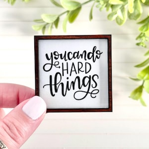 May include: Small square wooden sign with the phrase "you can do hard things" in black script on a white background. The sign has a dark brown frame and is held by a person.