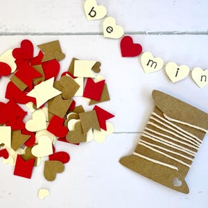 May include: A collection of red, brown, and white paper hearts, squares, and flags, along with a spool of twine. The twine is wrapped around a brown cardboard spool with a heart-shaped cutout. A banner with the words "be mine" is strung with white hearts.