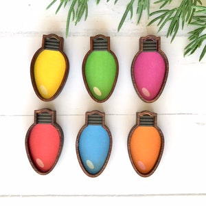 May include: Six wooden Christmas light ornaments in various colors, including yellow, green, pink, red, blue, and orange. The ornaments are shaped like light bulbs and have a brown wooden frame.