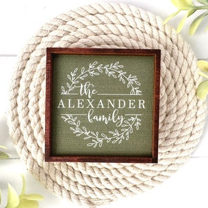 May include: A green wooden sign with a brown frame, featuring a white wreath with the text "the Alexander family" in a cursive font.