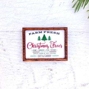 May include: A rectangular wooden sign with the text "FARM FRESH Christmas Trees" in white and red. The sign also includes the text "PINE &bull; SPRUCE &bull; FIR &bull; CEDAR" and "APPLE CIDER & HOT COCOA".