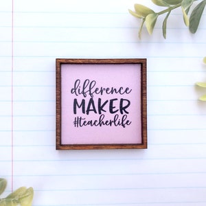 May include: A small wooden sign with a light pink background and a dark brown frame. The sign says "difference MAKER #teacherlife" in black text.