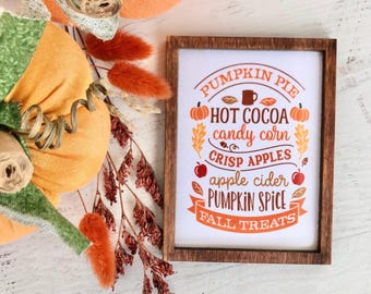 May include: A rectangular wooden sign with white background and orange and brown text reading PUMPKIN PIE, HOT COCOA, candy corn, CRISP APPLES, apple cider, PUMPKIN SPICE, and FALL TREATS, decorated with small pumpkin and leaf icons.