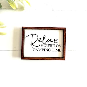 May include: A rectangular wooden sign with a dark brown frame and a white background. The sign features black text that reads "Relax YOU'RE ON CAMPING TIME" in a combination of script and block lettering centered on the white surface.