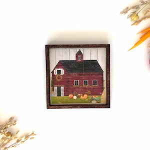 May include: A framed illustration of a red barn with a white door and windows, surrounded by pumpkins and gourds. The barn has a black roof and a white picket fence in the background.