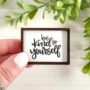 May include: A small, rectangular wooden sign with the phrase "be kind to yourself" in black script. The sign has a white background and a dark brown frame. Green foliage is visible in the background.
