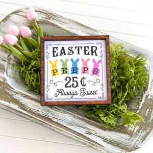 May include: A wooden sign with a white background and brown frame. The sign says "Easter Peeps 25¢ Always Sweet" with a graphic of five colorful Peeps. The sign is sitting in a whitewashed wooden bowl with green moss.