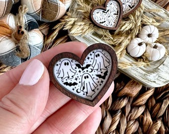 May include: A heart-shaped wooden pin featuring a white background with a black graphic print of ghosts, bats, stars, and crescent moons. The dark brown wood frame surrounds the central design, which is held in a hand with light pink nails.