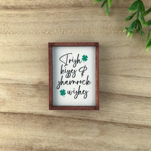 May include: A wooden sign with a white background and brown frame. The sign has green shamrock designs and the text "Irish kisses & shamrock wishes".