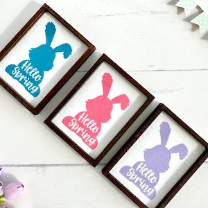 May include: Three wooden signs with a bunny silhouette and the text "Hello Spring" in white. The bunnies are in shades of blue, pink, and purple.