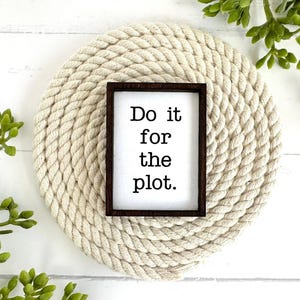 May include: A rectangular sign with a dark brown wooden frame and a white background featuring the black text 'Do it for the plot.' The sign is centered on a circular, coiled beige rope mat, surrounded by green artificial foliage.