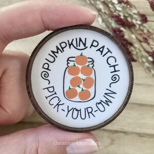 May include: A round wooden sign with a white background and brown trim. The sign says "Pumpkin Patch Pick-Your-Own" in a black script font. A jar of orange pumpkins is illustrated in the center of the sign.