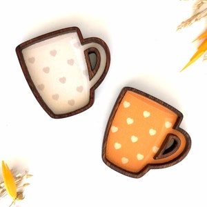 May include: Two wooden mugs with a heart pattern. One mug is white with light brown hearts and the other is orange with white hearts.