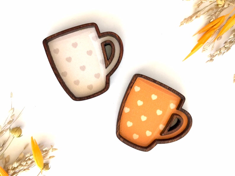 May include: Two wooden mugs with a heart pattern. One mug is white with light brown hearts and the other is orange with white hearts.