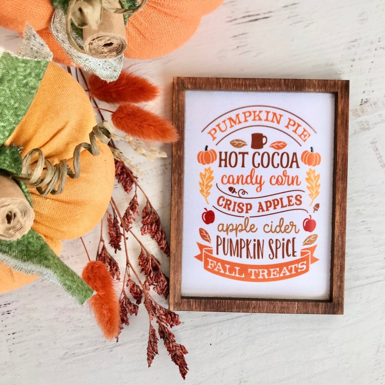 Może przedstawiać: Drewniany znak z białym tłem i brązowymi literami. Znak zawiera napis "Pumpkin Pie, Hot Cocoa, Candy Corn, Crisp Apples, Apple Cider, Pumpkin Spice, Fall Treats". Znak jest otoczony jesiennymi ozdobami, w tym dyniami i jesiennymi liśćmi.