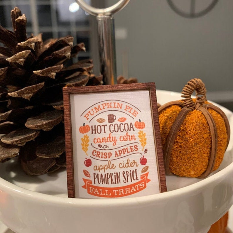 Może przedstawiać: Drewniany znak z brązową ramą i białym tłem. Znak zawiera napis "Pumpkin Pie, Hot Cocoa, Candy Corn, Crisp Apples, Apple Cider, Pumpkin Spice, Fall Treats". Są małe ilustracje dyni, liści i kubka gorącej czekolady.