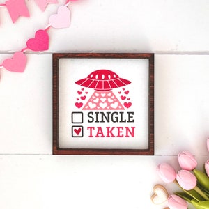 May include: A wooden sign with a red UFO and hearts, with the text "SINGLE" and "TAKEN" with a checkmark next to "TAKEN".