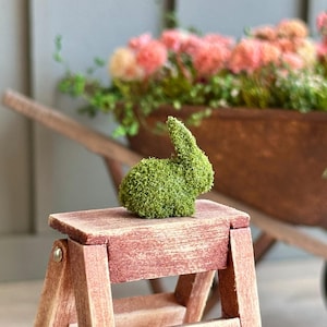 May include: A small green moss bunny figurine sits on a wooden step stool. The stool is made of weathered wood and has a reddish brown stain. The bunny is sitting on the top step of the stool.