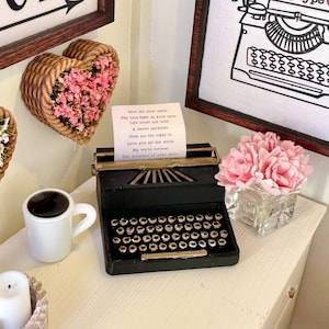 May include: A miniature black typewriter with a paper insert that reads "Give me your smile, the love sighs in your eyes, Life smells to be wild, A fairer paradise, Give you the right to love you all the while, My world forever, The treasure of your smile". The typewriter is on a white surface with a cup of coffee, a vase of pink flowers, and a heart-shaped wicker basket filled with pink flowers.