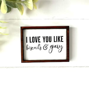 May include: A wooden sign with a brown frame and white background. The sign reads "I love you like biscuits & gravy".
