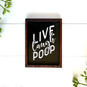 May include: A black wooden sign with a white inscription that reads "LIVE laugh POOP" in a decorative font. The sign has a dark brown wood frame.