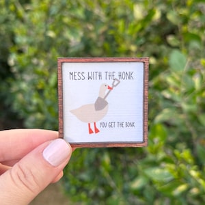 May include: A small, square wooden sign with the text "MESS WITH THE HONK YOU GET THE BONK." The sign features a cartoon goose holding a shovel. The goose is beige with red feet and a grey scarf.