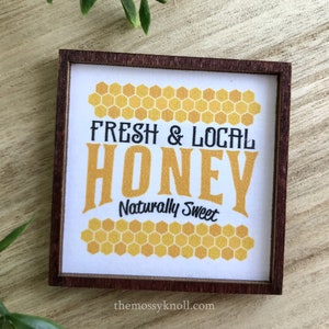 May include: A wooden sign with a brown frame and a white background. The sign says "Fresh & Local Honey Naturally Sweet" in black text with a yellow honeycomb pattern around the text.