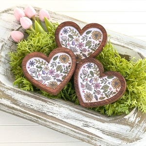 May include: Three wooden heart-shaped ornaments with a floral pattern in shades of pink, purple, yellow, and green. The hearts are nestled in a whitewashed wooden bowl filled with green moss.