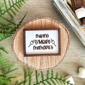 May include: A wooden sign with the text "Making S'more Memories" in a brown wood frame. The sign is sitting on a wooden slice.
