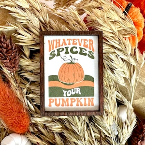 May include: A wooden sign with the text "Whatever Spices Your Pumpkin" in a retro font. The sign features a stylized orange pumpkin with a green stem and is surrounded by a green and orange border.