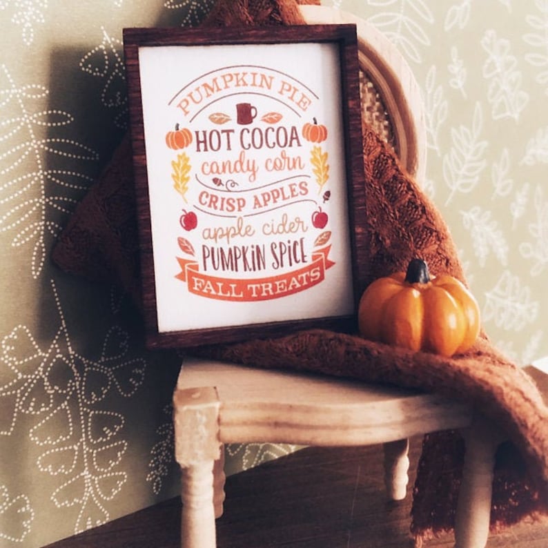 Może przedstawiać: Ramka z napisem "Pumpkin Pie, Hot Cocoa, Candy Corn, Crisp Apples, Apple Cider, Pumpkin Spice, Fall Treats". Ramka stoi na małym drewnianym krześle z brązowym kocem i małą pomarańczową dynią.
